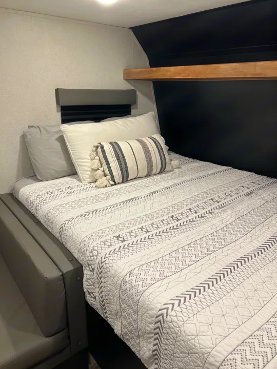 Compact RV rental bedroom with a gray patterned quilt, pillows, and a wood shelf for cozy travel.. Keystone Springdale 2022