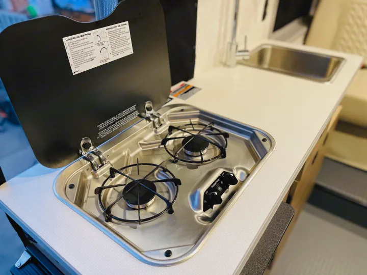 RV rental kitchen: a two-burner stove, sink, and faucet on a countertop. Instructions for use are on the lid.. Winnebago Winnebago 2023