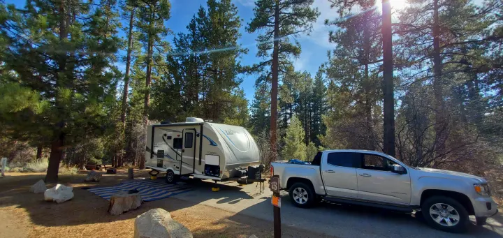 An RV rental travel trailer is set up at a sunny campsite with a silver pickup truck parked nearby, surrounded by tall pine trees.. Coachmen Other 2019