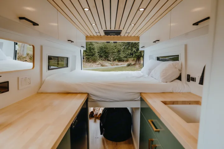 Modern **rv rental** camper van interior: elevated bed, wood ceiling/counters, forest view. Ideal for road trip comfort.. Ram Promaster 2500 2021