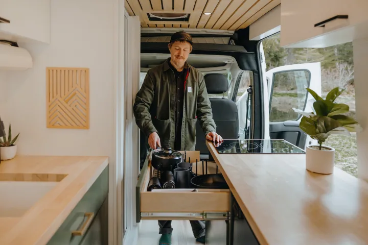 A man in an RV rental kitchen pulls cookware from a drawer, ready to cook in the modern camper van amidst nature.. Ram Promaster 2500 2021
