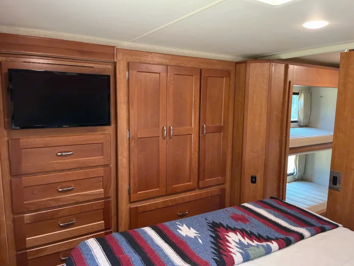 Interior of an **rv rental** showing wood cabinetry, a mounted TV, bed with a patterned blanket, and visible bunk beds. Ideal for family travel.. Winnebago Minnie Winnie 2017