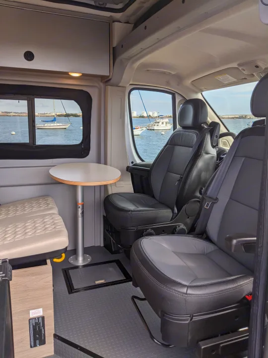 RV rental interior with swivel seats, a table, and windows overlooking a bay with boats.. Winnebago Solis 2023