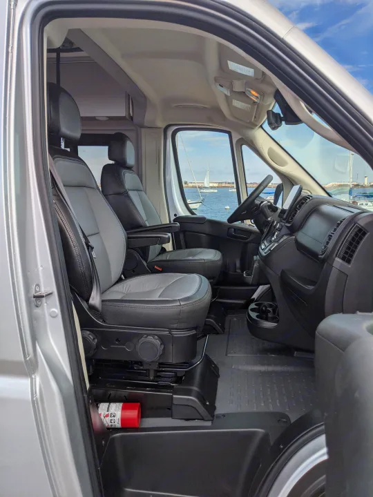 RV rental van interior: view from open passenger door of seats, dashboard. Ocean with sailboats outside. Fire extinguisher.. Winnebago Solis 2023