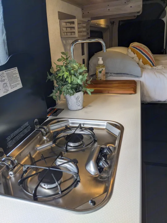Modern RV rental: a compact kitchen setup with a stainless steel two-burner stove, sink, and a comfortable bed.. Winnebago Solis 2023
