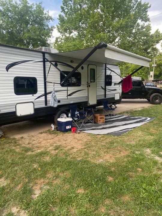 RV rental campsite scene: A travel trailer with an awning extended, chairs, a cooler, and a mat on the ground. A black Jeep is parked behind.. Avenger ATI 2015