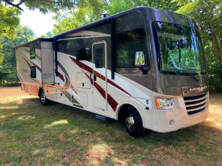 This RV rental shows a large Class A motorhome, extended with slide-outs, parked on grass beneath trees.. Forest River Coachmen Mirada 2019