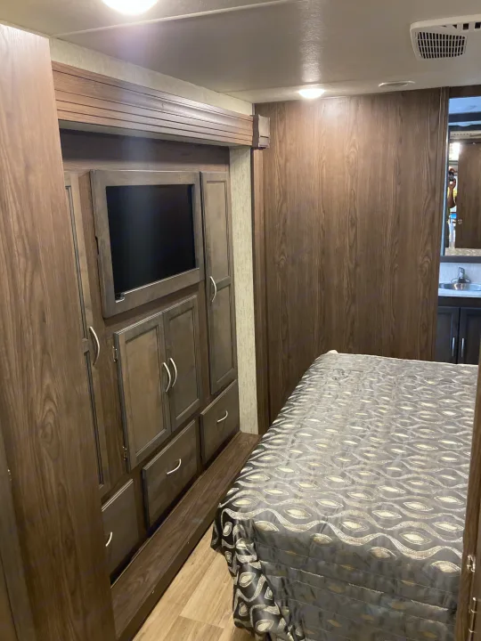 Dark wood RV rental bedroom with built-in TV, storage, and bed. Bathroom sink visible in mirror.. Forest River Coachmen Mirada 2019