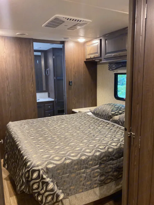 RV rental interior: A cozy bedroom with a patterned bed and pillows, leading to a bathroom. Features overhead AC and storage.. Forest River Coachmen Mirada 2019
