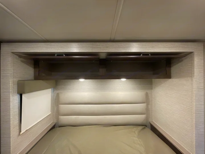 Inside an RV rental: a modern bedroom with a comfy bed, padded headboard, open overhead cabinets, and reading lights.. Winnebago Sunstar 2022