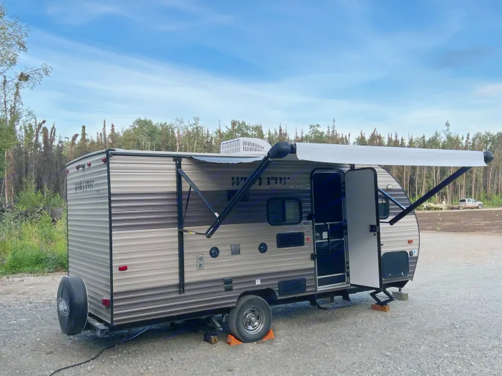 A grey Wolf Pup travel trailer, ideal for an RV rental, is parked on gravel with its awning extended, surrounded by trees under a blue sky.. Forest River Cherokee Wolf Pup 2018