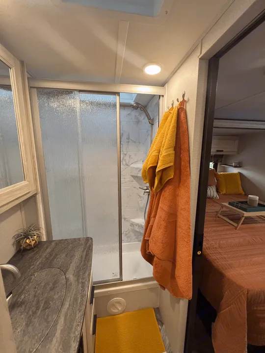 Inside a modern RV rental: a bathroom with a glass shower and vanity, leading to a cozy bedroom with an orange bedspread.. Forest River Sunseeker 2023