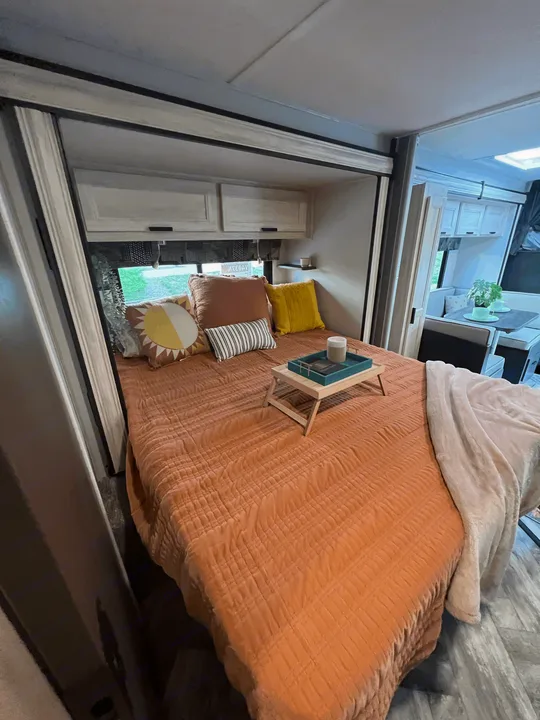 Inside a modern RV rental: a comfy bed with orange quilt, pillows, and a breakfast tray. Dinette visible.. Forest River Sunseeker 2023