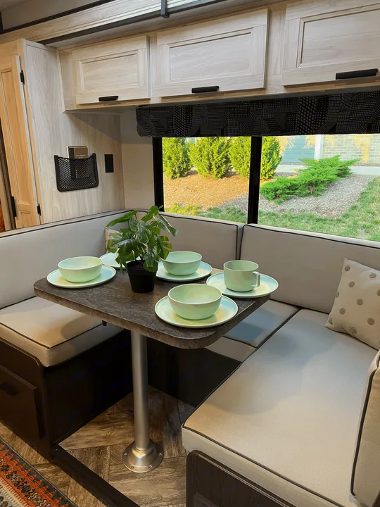 Spacious RV rental dinette with light grey booth seating, a dark table, mint green dishes, and a view of green foliage outside.. Forest River Sunseeker 2023