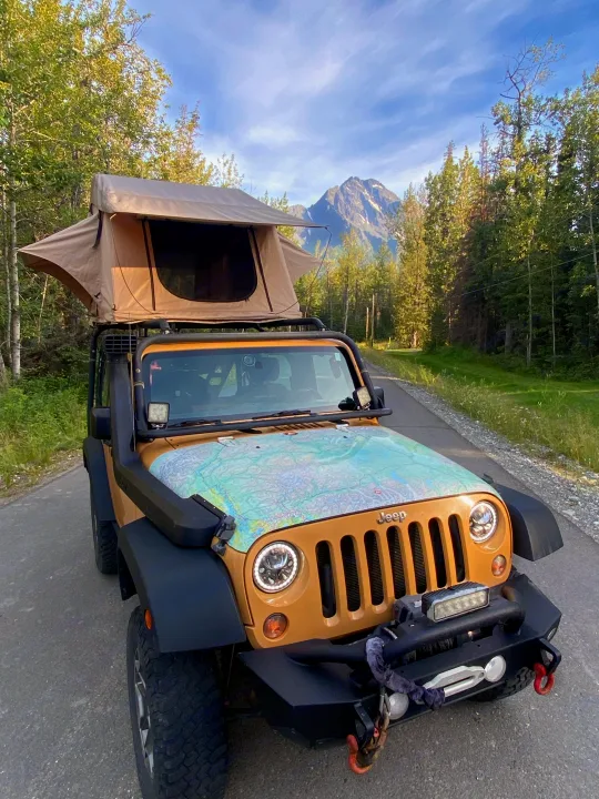 Tan Jeep Wrangler with rooftop tent and hood map, parked on a forest road with mountain views. A great RV rental alternative!. Jeep Rubicon 2014