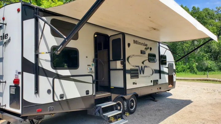 An RV rental: Winnebago Minnie Plus travel trailer with awning extended, steps down. Parked on gravel at a sunny campsite.. Winnebago Winnebago Minni Plus 27BHSS 2018