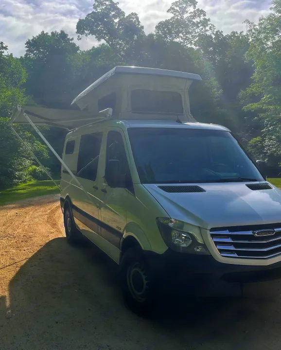 This white Freightliner pop-top rv rental, with its side awning out, is parked on a dirt road in a sunny forest.. Freightliner Sprinter 2015