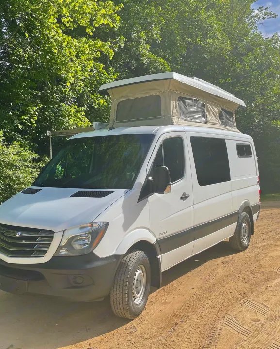 White camper van with a raised pop-top tent, parked on a dirt road by trees. Perfect for your next RV rental adventure!. Freightliner Sprinter 2015