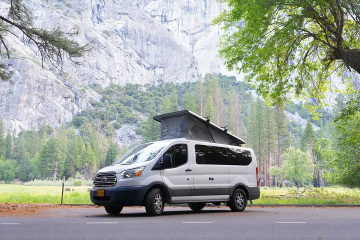 Pop-top RV rental camper van parked in Yosemite Valley, backed by towering granite mountains and lush forests.. Ford transit 2016