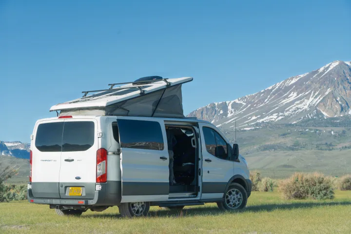 White pop-top camper van, an rv rental, on grass before mountains. Ready for your next outdoor adventure.. Ford transit 2016