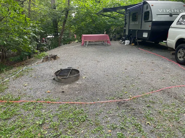 An RV rental travel trailer is parked on a gravel campsite with a picnic table and fire pit, surrounded by trees.. Other 1906st Saga 2021