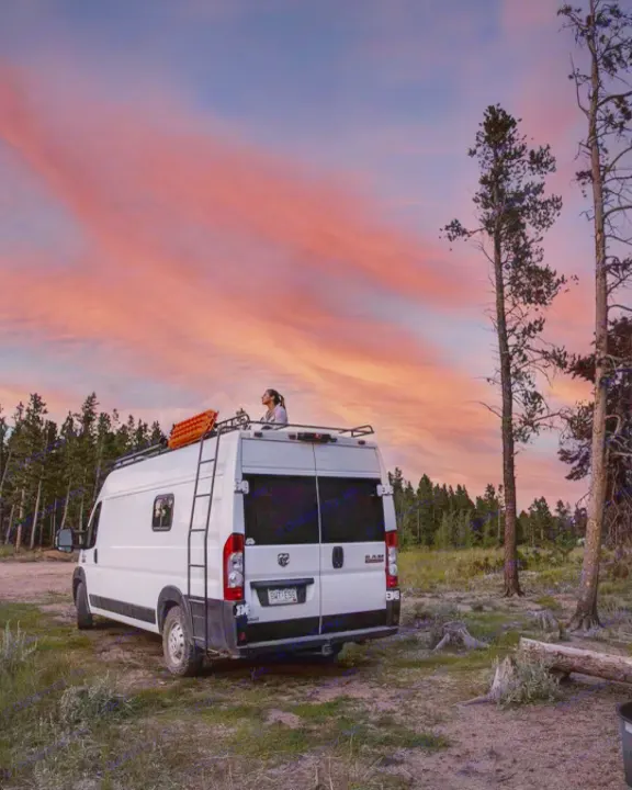 Serene sunset from an rv rental. A person watches the vibrant sky from atop a white camper van, parked amidst tall pine trees.. Other Other 2021