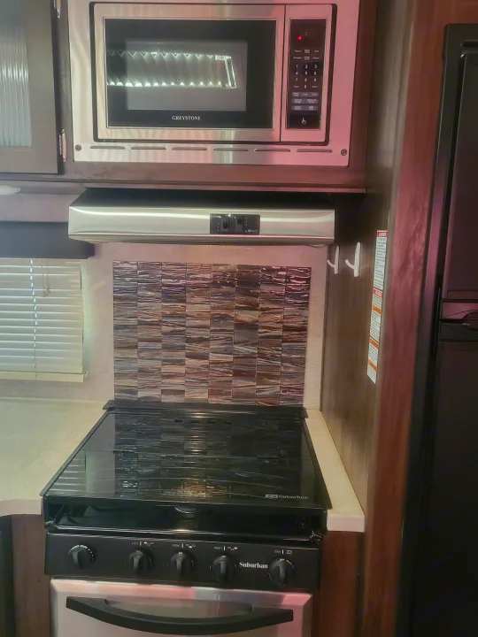 This RV rental kitchen features a Greystone microwave, Suburban oven/range, range hood, and a decorative tiled backsplash.. Forest River Cherokee Grey Wolf 2018