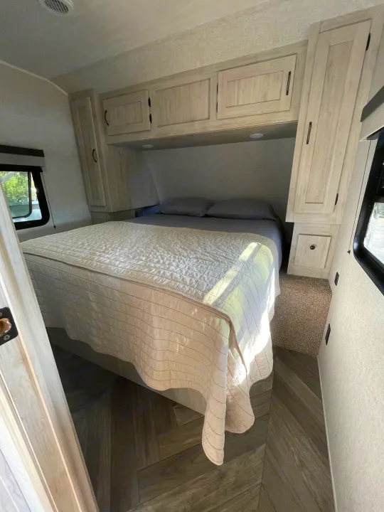 RV rental bedroom interior featuring light wood cabinets, a comfortable bed, and diagonal wood-look flooring.. East to West Other 2022