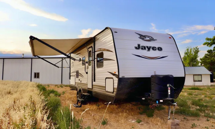 A white Jayco RV rental travel trailer is parked with its awning out, surrounded by dry grass and buildings.. Jayco Jay Flight SLX 2018