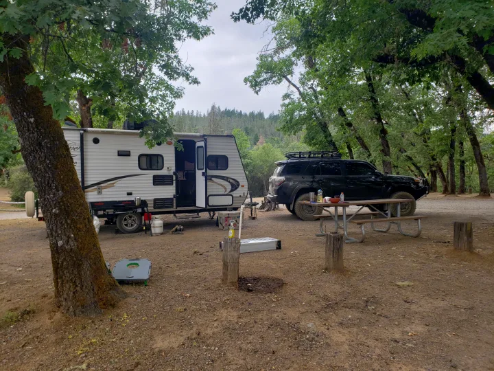 Serene campsite featuring an RV rental, a black SUV, picnic table, and various camping gear under leafy trees.. Clipper Clipper Trailer 2015