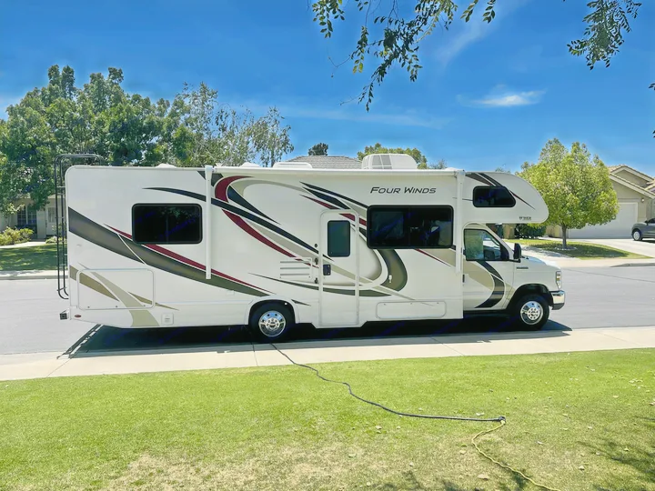 White Four Winds RV rental, a Class C motorhome, parked curbside on a sunny day. Hoses visible on the green lawn.. Thor Four Winds 2021