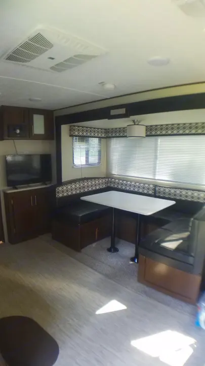 Inside an RV rental: a comfortable U-shaped dinette with table, TV area, windows with blinds, and AC unit overhead.. Avenger ATI 2018