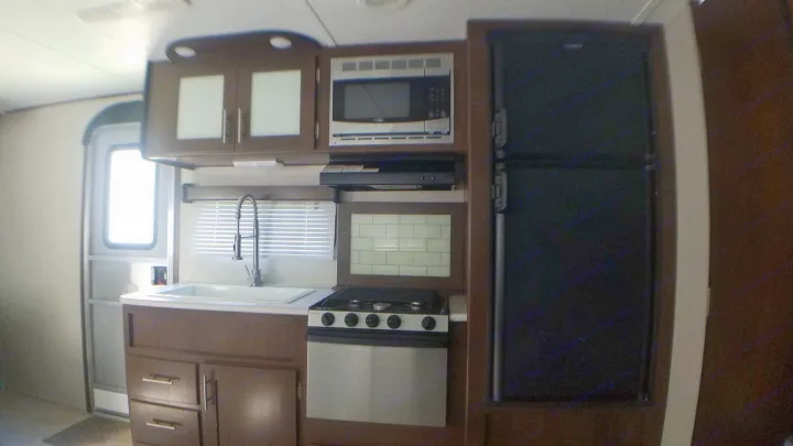 Spacious RV rental kitchen featuring a sink, stove, microwave, and refrigerator. Dark wood cabinets & blinds on window.. Avenger ATI 2018