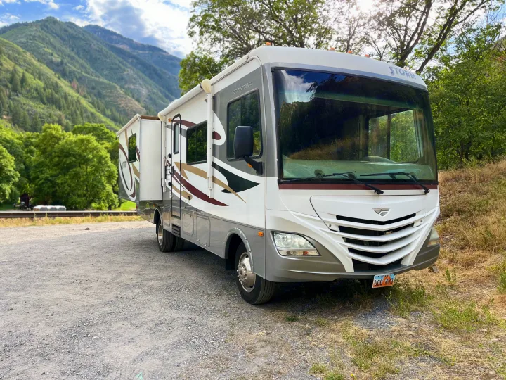 White & grey RV rental with extended slide-out, parked on gravel against lush green mountains. Perfect for scenic travel.. Fleetwood Storm 32BH 2013