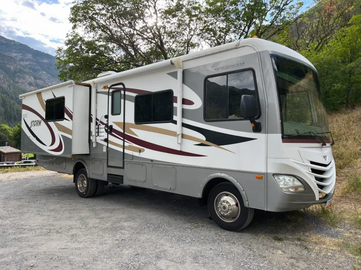 This spacious Fleetwood Storm RV rental, with a slide-out extended, is parked on gravel amidst scenic mountains.. Fleetwood Storm 32BH 2013