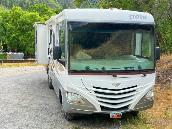 This white Storm RV rental, with a slide-out extended, is parked on a gravel lot amidst lush green scenery.. Fleetwood Storm 32BH 2013