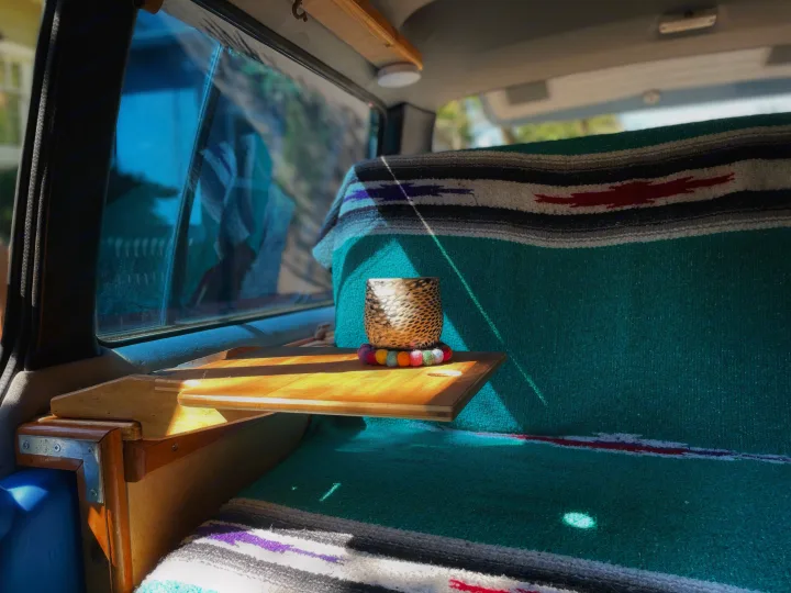 Enjoy the cozy RV rental interior, featuring a wooden shelf, patterned teal blanket, and a cup in sunlight.. Toyota Hiace 1985