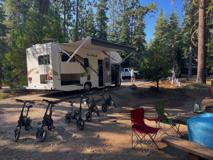 A Four Winds RV rental is parked at a shaded campground site, with four electric bikes, chairs, and a picnic table, under tall trees.. Thor Four Winds 2019