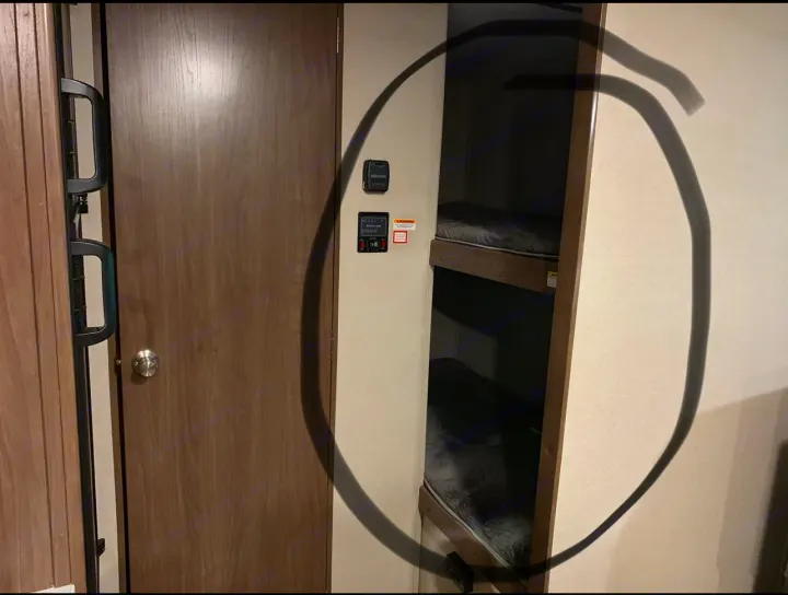 RV rental interior: a light wood door, wall controls, and cozy bunk beds with dark mattresses, framed by a dark circle.. Keystone RV Hideout 2019
