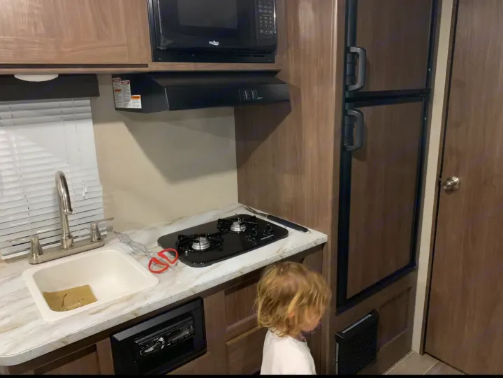 RV rental interior features a compact kitchen with sink, stovetop, microwave, fridge, and a child exploring.. Keystone RV Hideout 2019