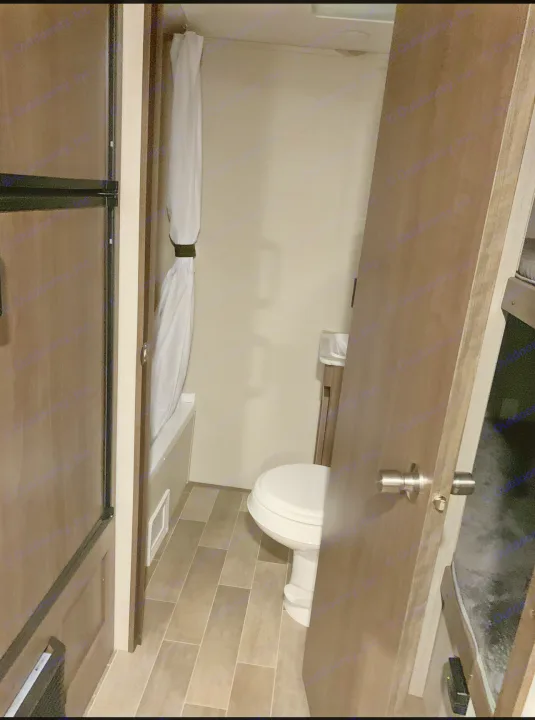 Compact RV rental bathroom with toilet, shower/tub, and tiled floor, adjacent to bunk beds and a refrigerator.. Keystone RV Hideout 2019