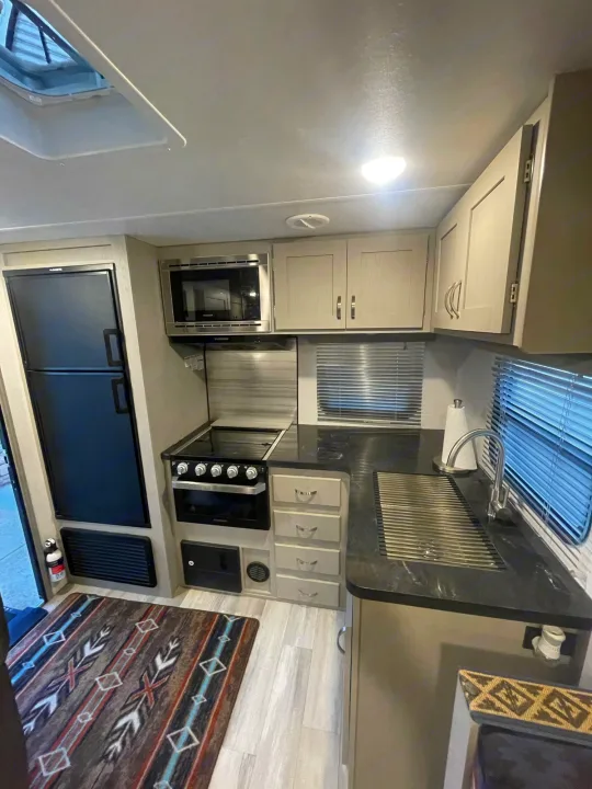 Inside an RV rental, this compact kitchen boasts a fridge, microwave, stove, sink with drain rack, light cabinets, and dark counters.. Prime Time Tracer 2021
