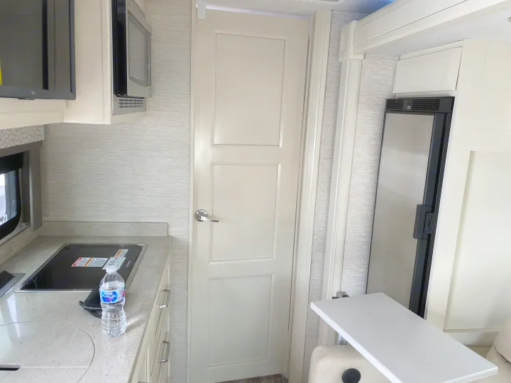 Compact RV rental interior: kitchen with stovetop, microwave, refrigerator, a door, and a side table.. Tiffin Motorhomes Wayfarer LW 2023