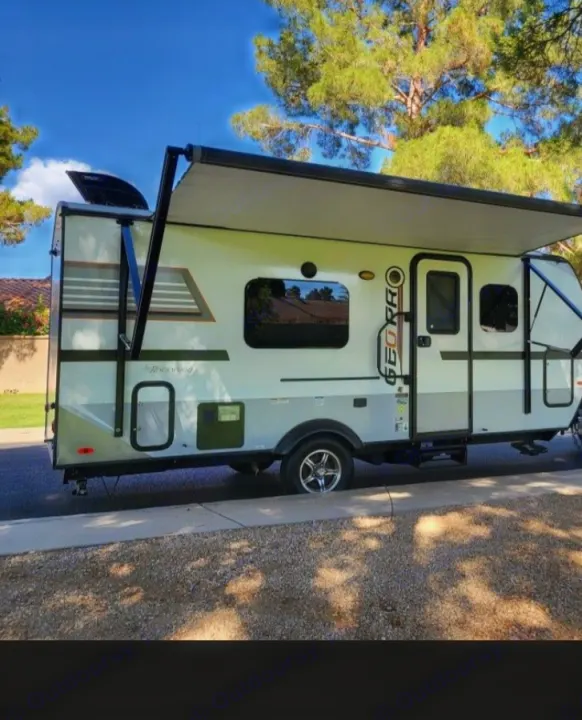 A light-colored compact RV rental, a travel trailer, parked on a street with its awning extended under a clear blue sky.. Forest River Rockwood Geo Pro 2018