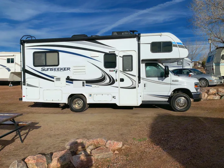 A white Sunseeker RV rental is parked at a sunny campsite, with other RVs and a car visible in the background.. Forest River Sunseeker 2019