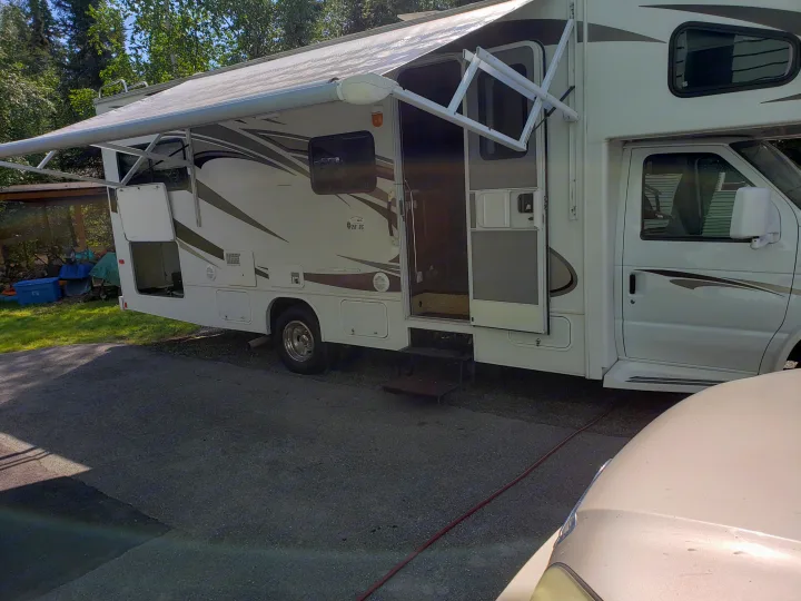 A white RV rental is parked outdoors with its awning extended and access panels open, showing available storage.. Jayco Greyhawk 2011