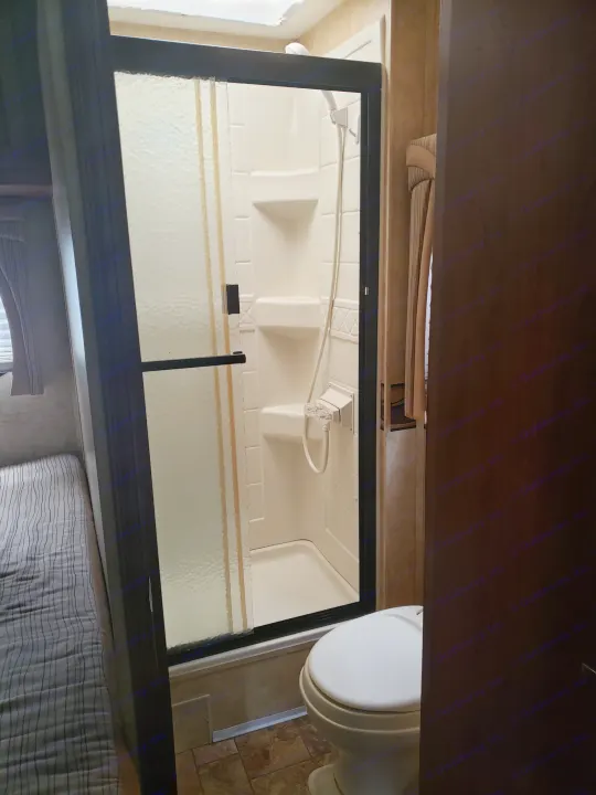 Inside an RV rental: a compact bathroom with a frosted glass shower enclosure and toilet, adjacent to a bed with striped linens.. Jayco Greyhawk 2011