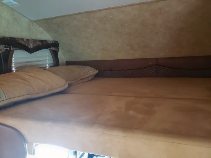 Cozy rv rental sleeping area with a comfortable bed, pillows, window blinds, and a handy storage net.. Jayco Greyhawk 2011