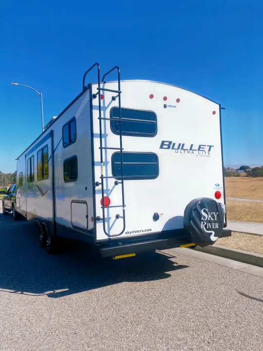 White Bullet travel trailer RV rental, featuring a rear ladder and Sky River spare tire cover, parked on a sunny day.. Keystone RV Bullet Ultra Lite 2022