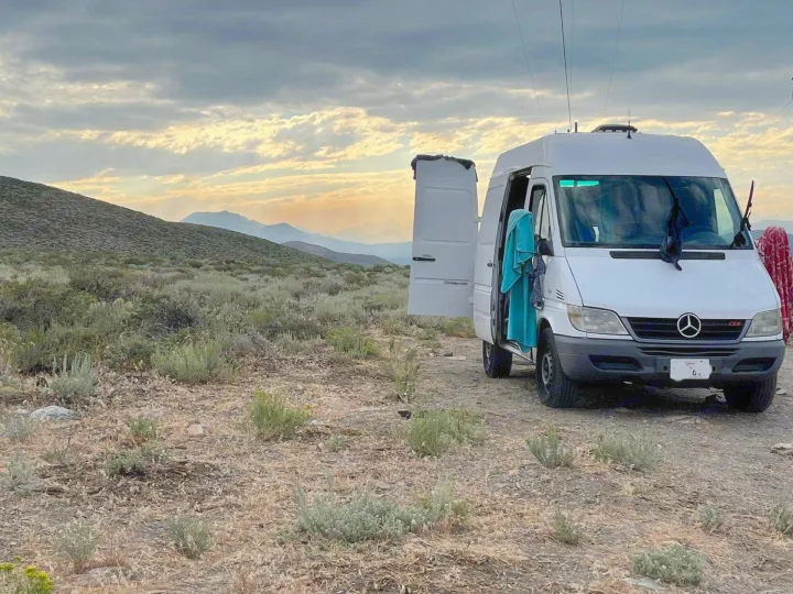 A white Sprinter camper van, potentially an RV rental, is parked in a vast, dry mountain landscape under a cloudy sunset sky.. Mercedes Sprinter 2006
